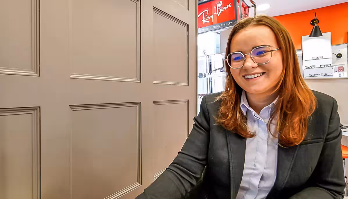 Smiling woman with glasses and brown hair wearing a black blazer and light blue shirt in an eyewear store with Ray-Ban signage visible.