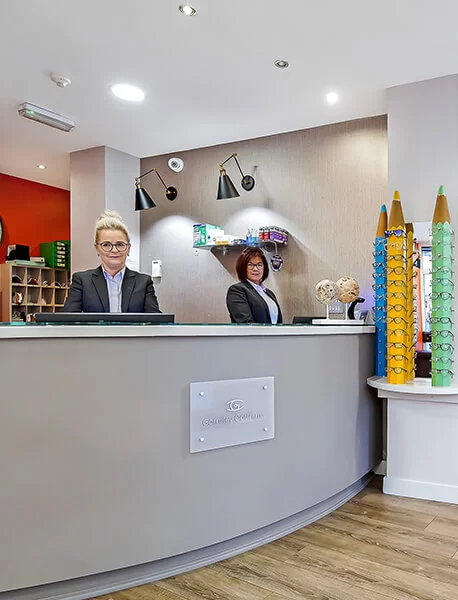 Two receptionists standing behind a gray curved reception desk in an optometry clinic with eyeglass displays visible on the right.