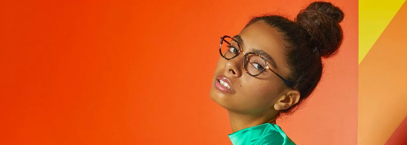 Woman with dark hair in a bun wearing tortoiseshell eyeglasses and a teal blouse against an orange and yellow background.