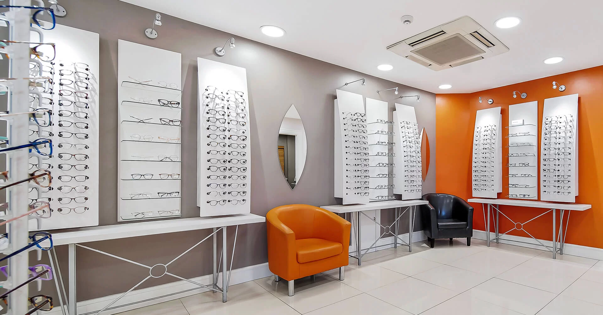Modern eyewear store interior with multiple white panels displaying various eyeglass frames, an orange chair, and a black chair against gray and orange walls.