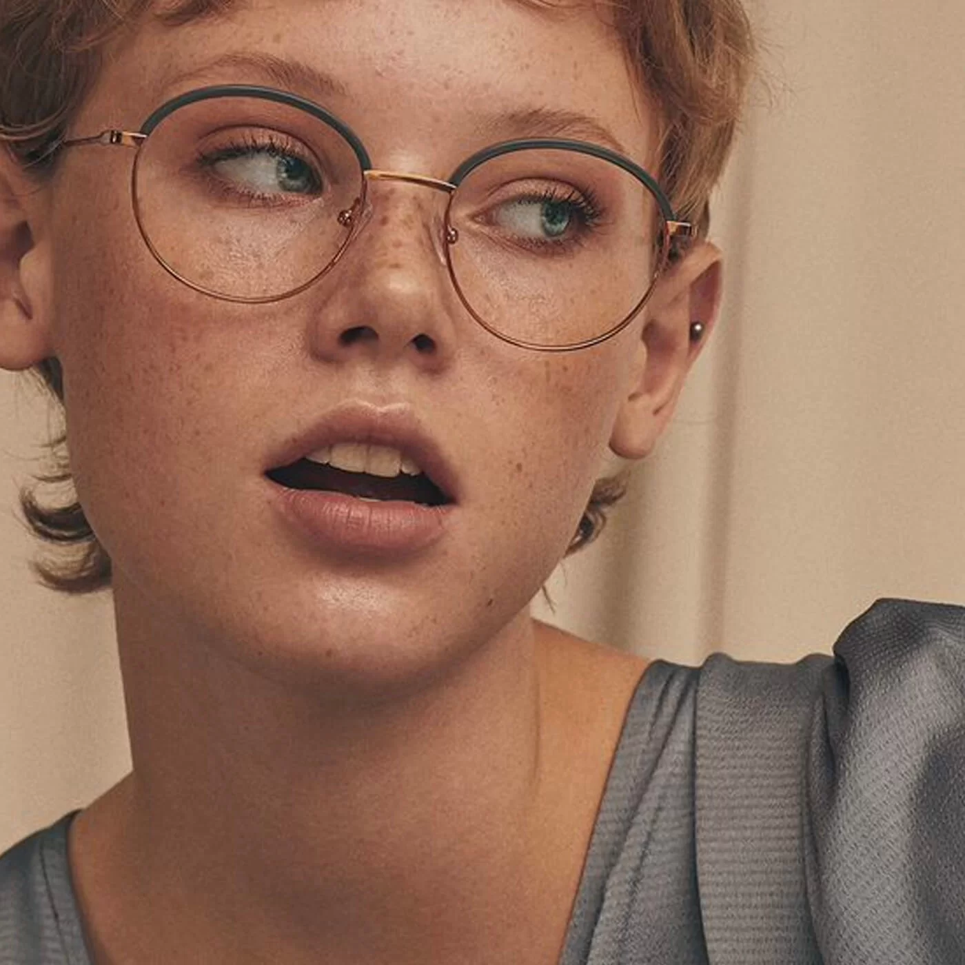 Close-up of a young person with short hair, freckles, wearing round glasses and a gray top, looking to the right.