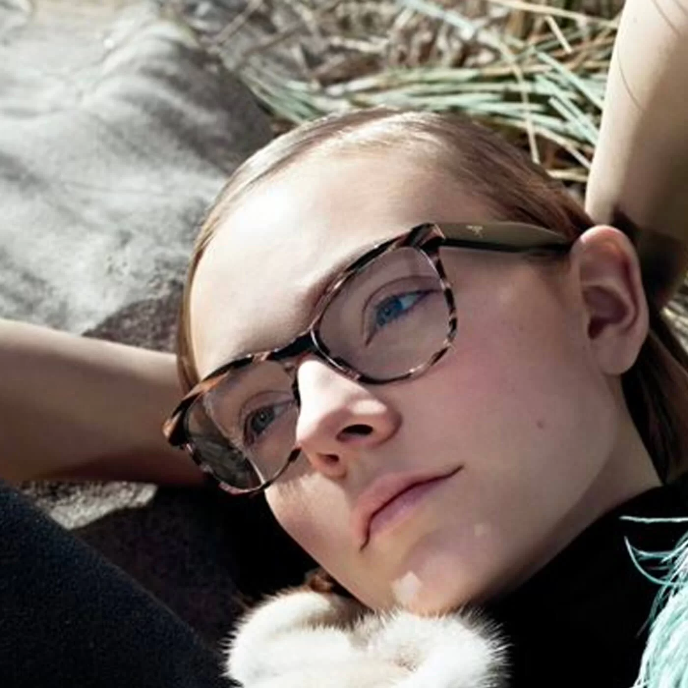 Close-up of a woman with light skin and blue eyes wearing tortoiseshell eyeglasses, resting her head on her arm outdoors.
