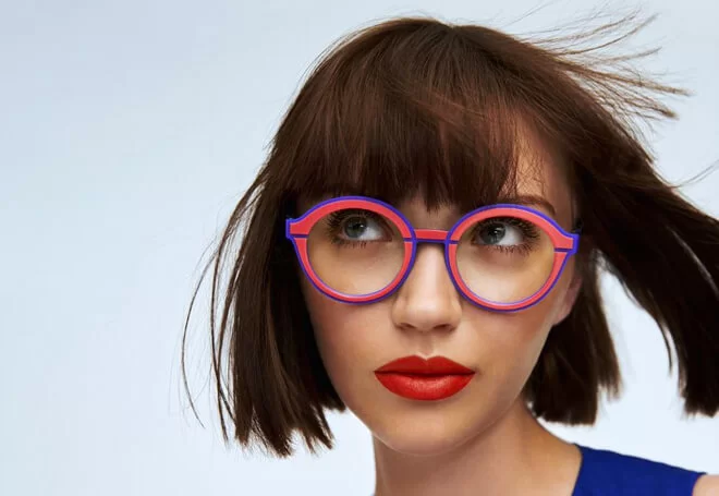 Young woman with short brown hair wearing round red and blue eyeglasses and bright red lipstick, looking slightly upward.