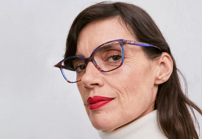 Close-up of a woman with long dark hair wearing red lipstick, tortoiseshell eyeglasses, and a white turtleneck sweater.