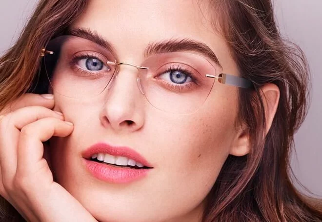 Close-up of a woman with blue eyes wearing thin-framed, rimless glasses and light pink lipstick, resting her chin on her hand.