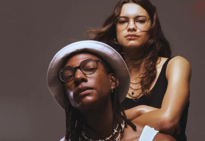 Portrait of two people wearing glasses, one with a white bucket hat and the other with long hair and a black tank top.