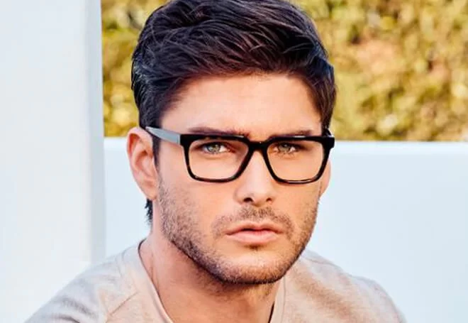 Man with dark hair and beard wearing black-rimmed glasses and a light-colored shirt, outdoors with blurred greenery background.