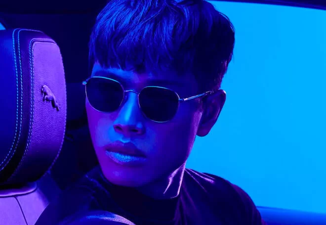 Man wearing round sunglasses and a dark shirt, sitting in a car with a horse emblem on the headrest, under blue lighting.