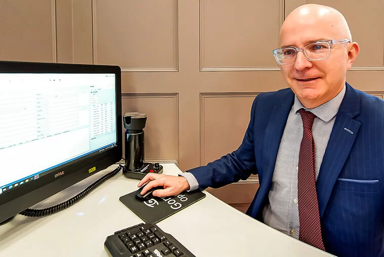 A smiling man in glasses and a suit seated at a desk using a computer mouse with a Dell monitor showing data.