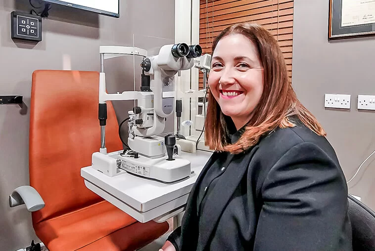 Smiling woman sitting next to eye examination equipment in an optometry clinic.