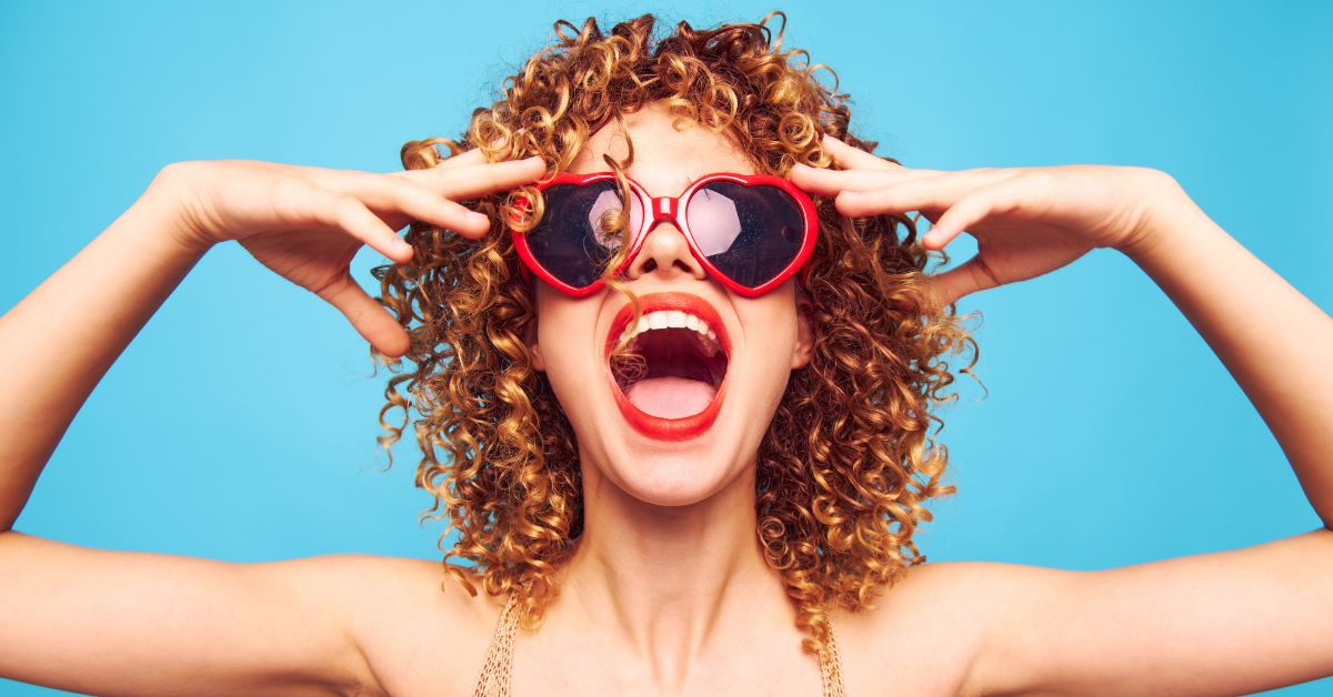 Excited woman with curly hair wearing bold red heart-shaped sunglasses.