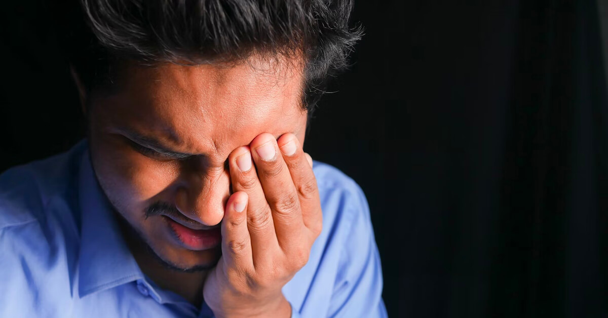 Man covering his eye in pain against a dark background.