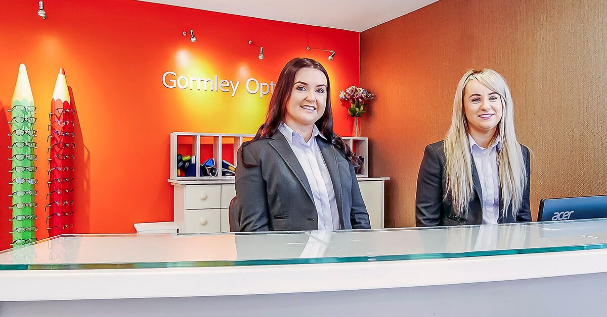 Two friendly staff members smiling at the Gormley Opticians reception desk.