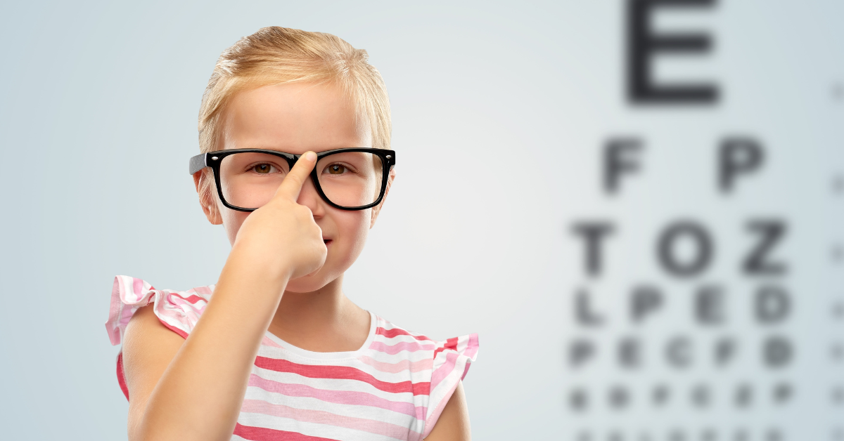 Young child pointing at black glasses in front of an eye chart.
