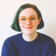 Young person with short green hair, orange glasses, and a navy blue sweater smiling against a plain white background.