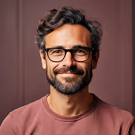 Smiling man with glasses, curly hair, and a beard wearing a muted pink shirt against a brown background.