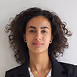 Portrait of a woman with curly black hair wearing a black blazer and white shirt, smiling slightly against a neutral background.