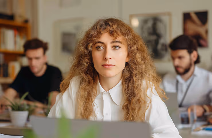 Young woman with curly hair in a white shirt working on a laptop in an office with two men in the background.