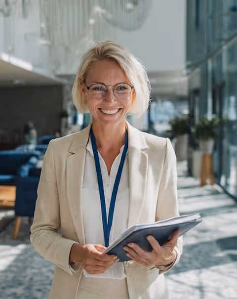 Smiling professional woman with blonde hair and glasses holding a clipboard in a modern office space.