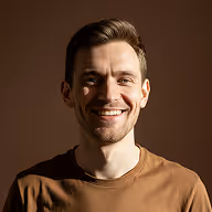 Smiling man with short brown hair wearing a brown shirt against a dark background.
