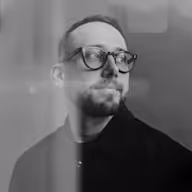 Man with glasses and facial hair wearing a dark shirt, looking slightly to the side in a grayscale photo.