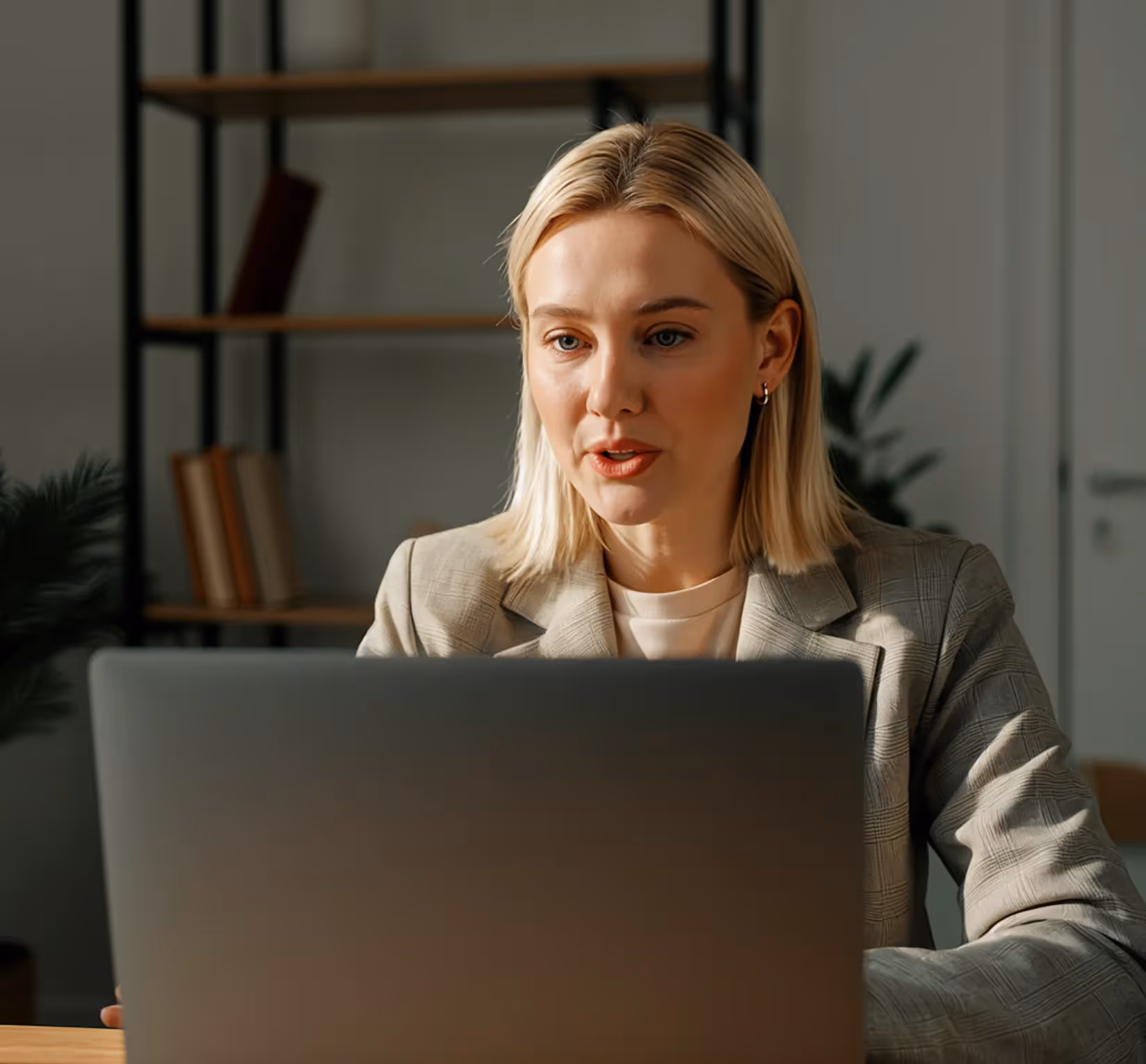 Blonde woman in a plaid blazer looking at a laptop screen in a softly lit office with bookshelves and plants in the background.