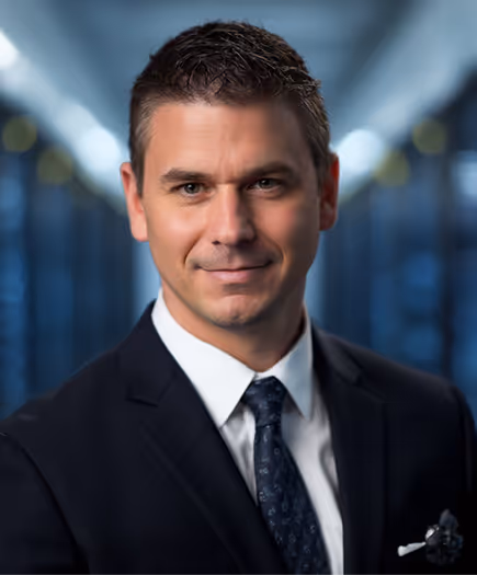 Smiling man in a dark suit and patterned tie with a blurred hallway background.