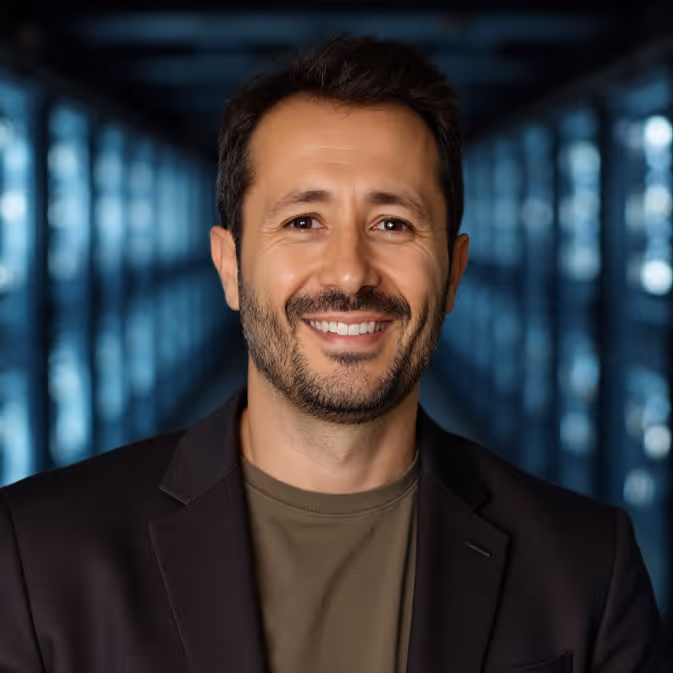 Smiling man with dark hair wearing a black blazer and olive shirt in a blurred corridor.