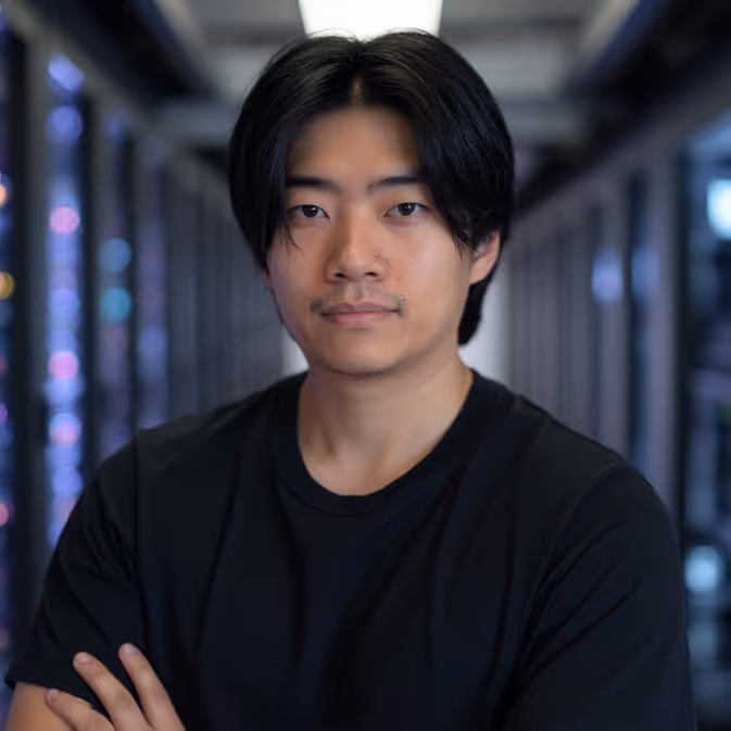 Young man with black hair wearing a black shirt standing with arms crossed in a tech server room.