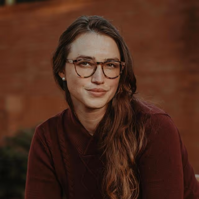 Young woman with long brown hair wearing tortoiseshell glasses and a maroon sweater, posing against a blurred brown background.