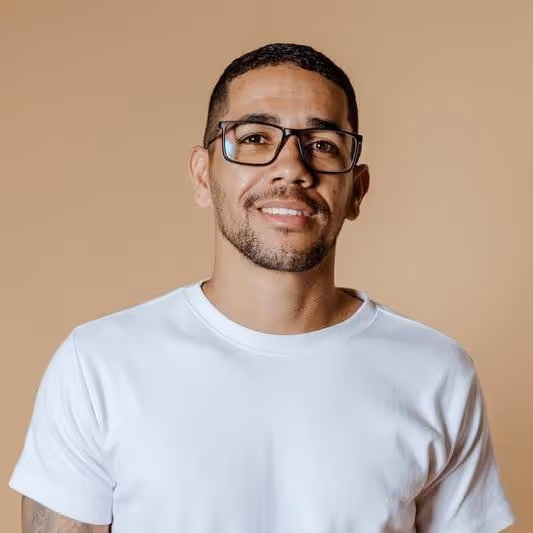 Smiling man with short hair, beard, and glasses wearing a white t-shirt against a beige background.