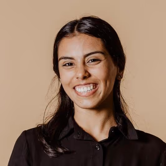 Young woman with long dark hair smiling against a plain beige background, wearing a black shirt.