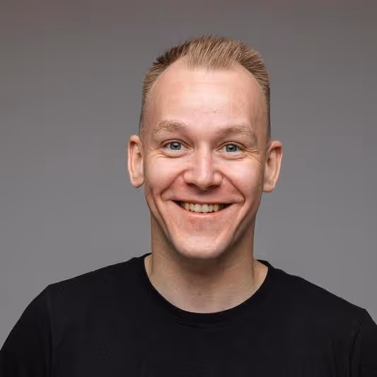 Smiling young man with short blond hair wearing a black shirt against a gray background.
