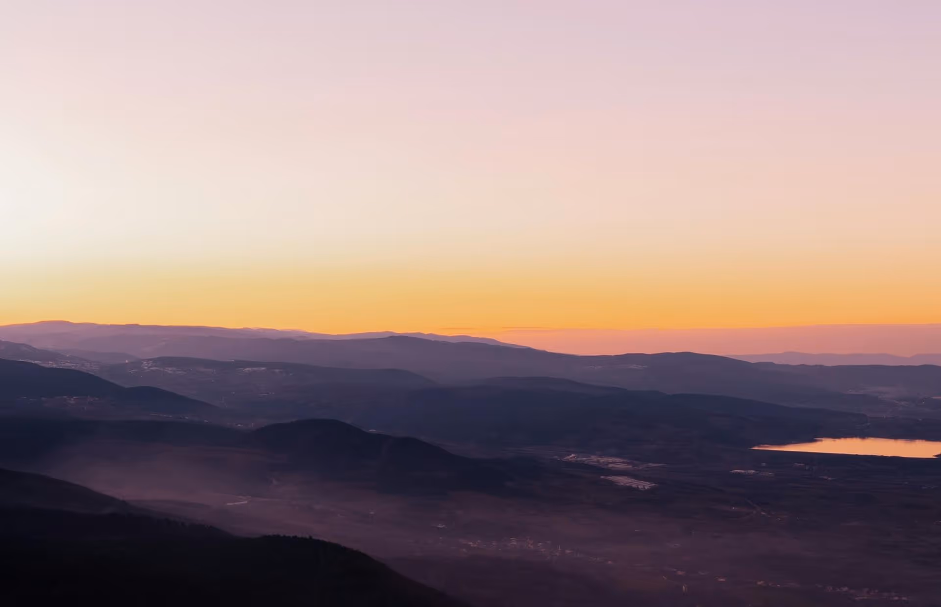 Layered mountain ranges at sunset with a calm lake reflecting orange light on the right.