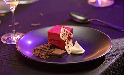 A purple plate topped with a piece of cake next to a glass of wine.
