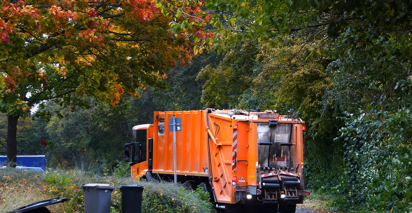 Orange garbage truck collecting waste in a leafy neighborhood during autumn with trash bins in the foreground.