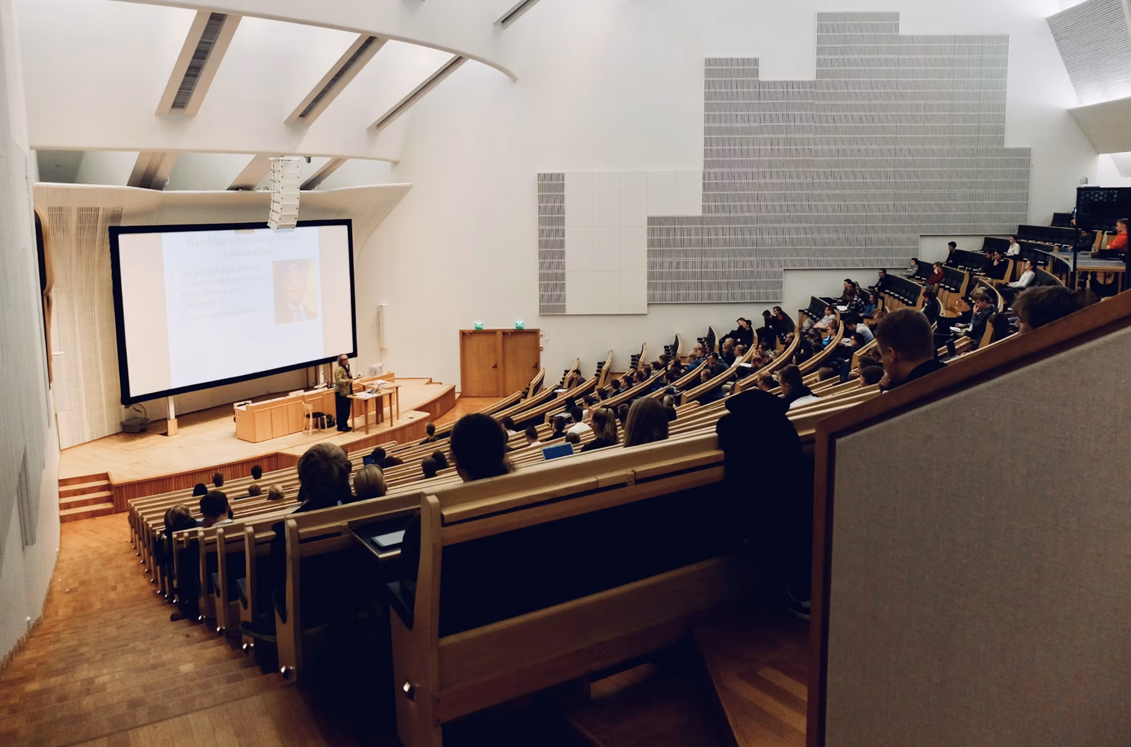 Interior of a modern lecture hall with tiered wooden seating filled with students facing a lecturer at a podium and a large projection screen displaying a presentation.