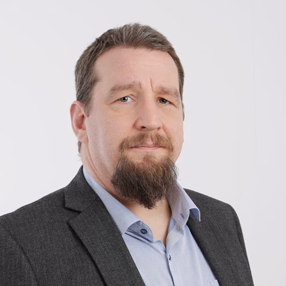 Portrait of a middle-aged man with a dark beard and mustache wearing a light blue shirt and dark gray suit jacket, looking at the camera with a neutral expression against a plain white background.