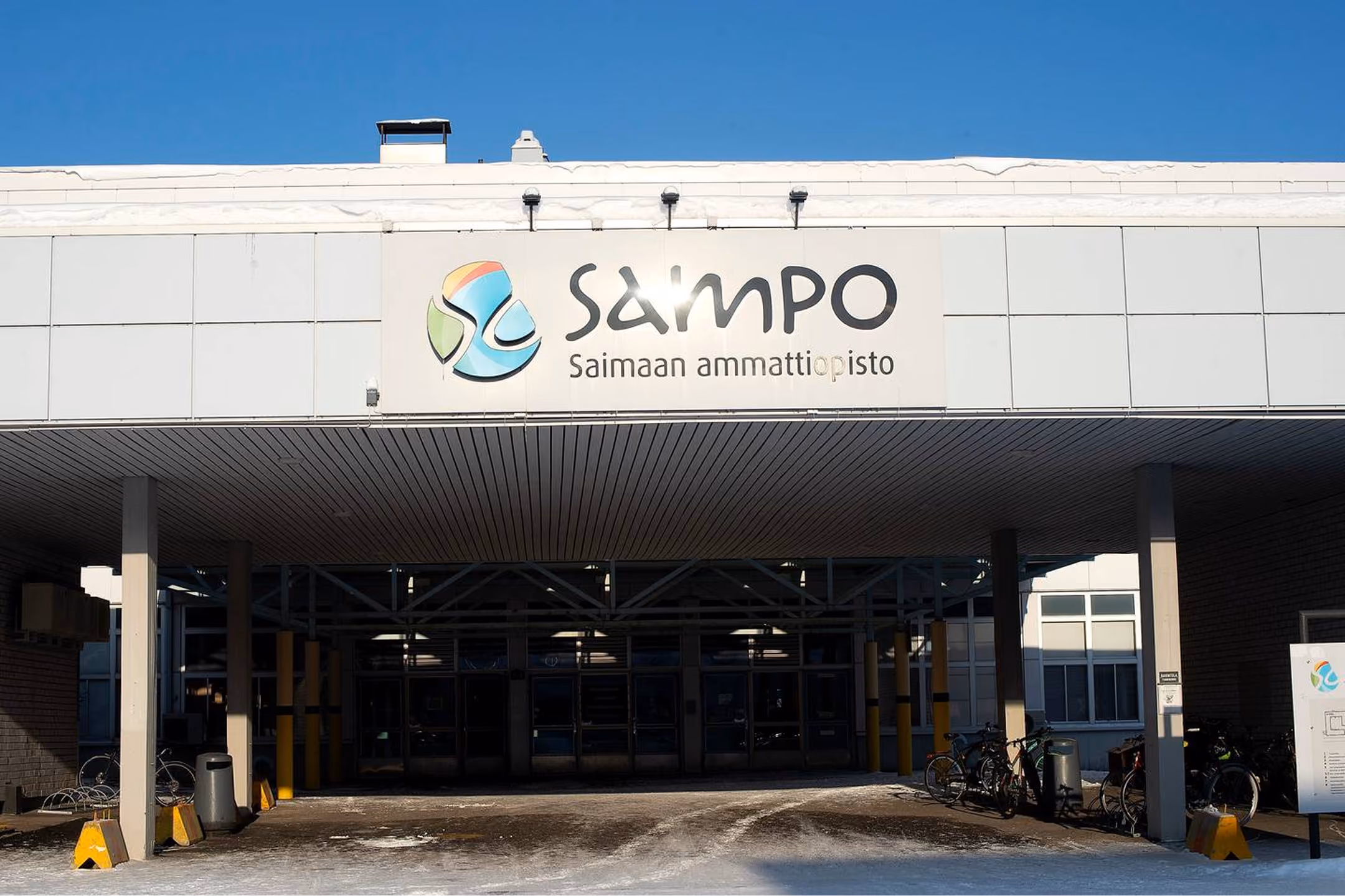 Entrance to a building with a sign reading 'Sampo Saimaan ammattiopisto,' with a colorful logo to the left of the text, under a clear blue sky.