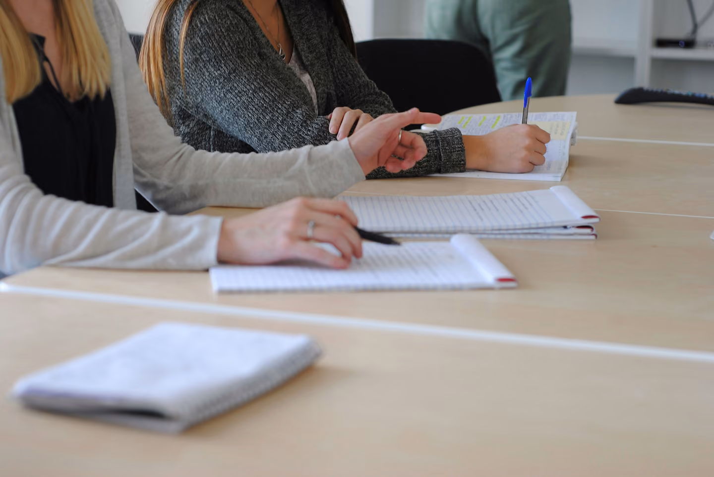 Two people sitting at a table with open notebooks, one person writing with a blue pen and the other holding a black pen and gesturing with their hand.