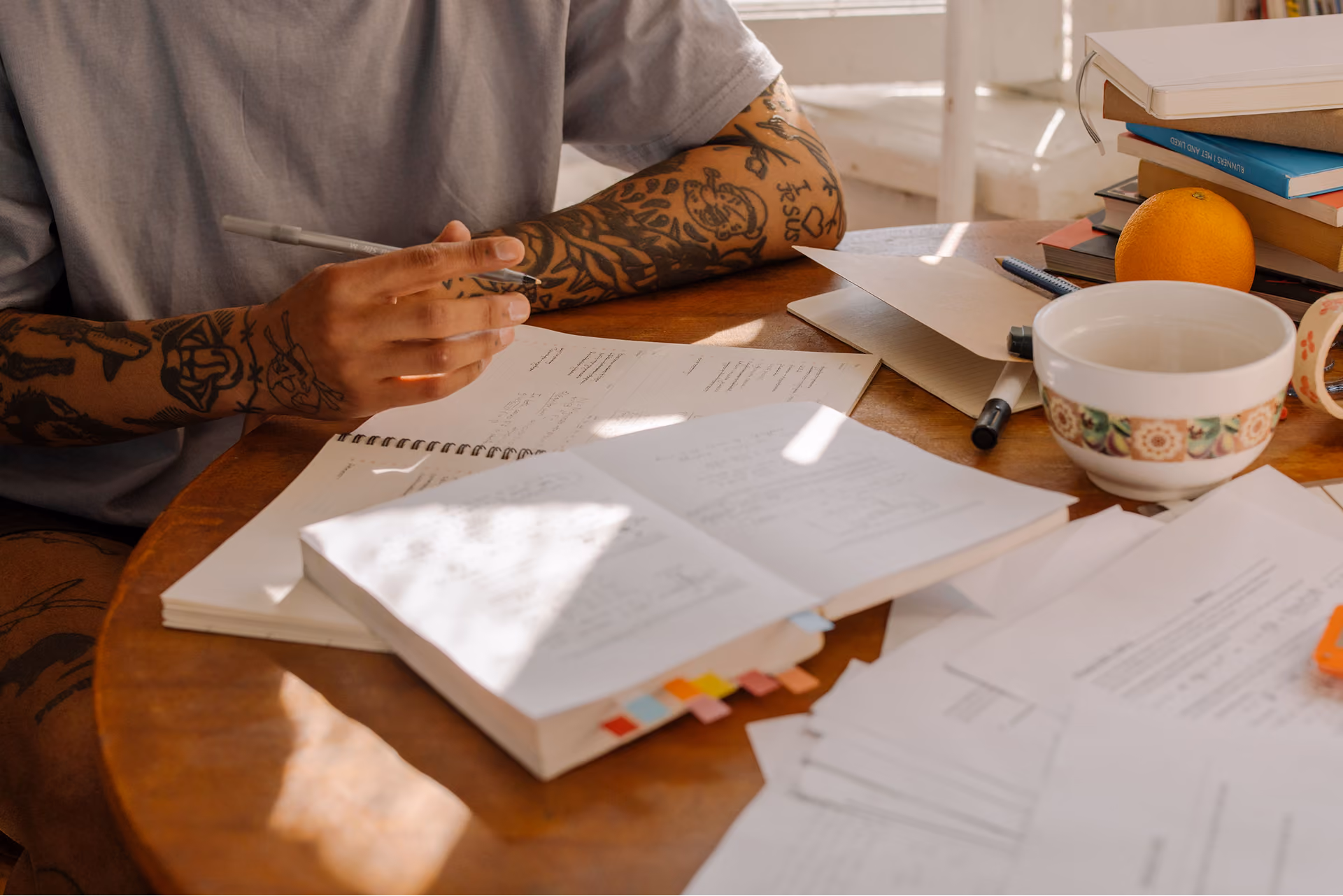 Tattooed person sitting at a wooden table filled with open notebooks, papers, pens, a bowl, an orange, and stacked books, studying or working.
