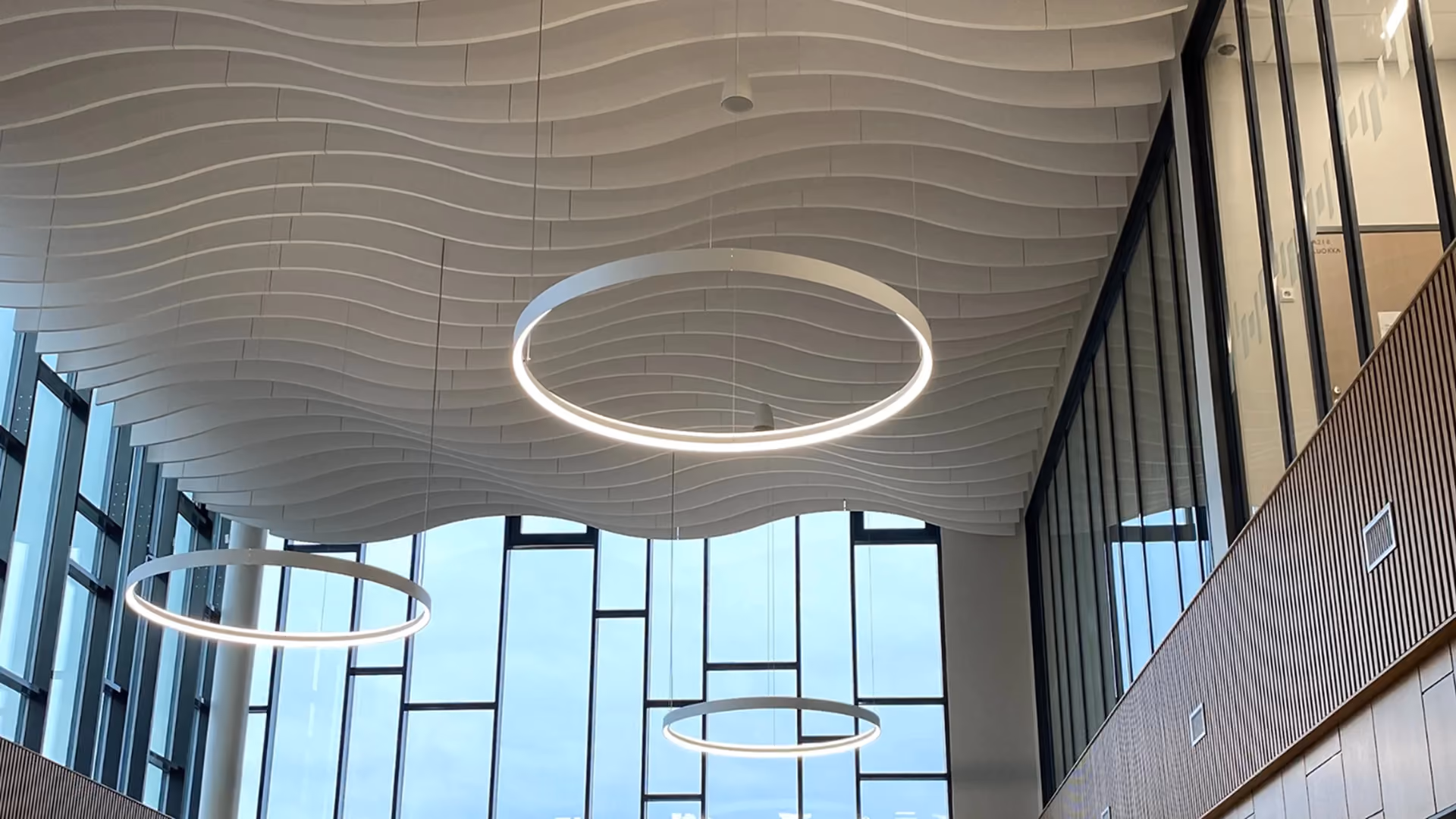 Interior ceiling with modern, wave-like white paneling and three large circular LED light fixtures hanging at different heights above large windows.