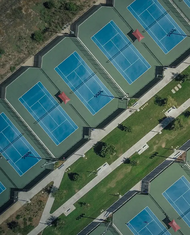 Aerial view of multiple outdoor tennis courts with blue playing surfaces separated by fences and a central grassy walkway with pathways and trees.