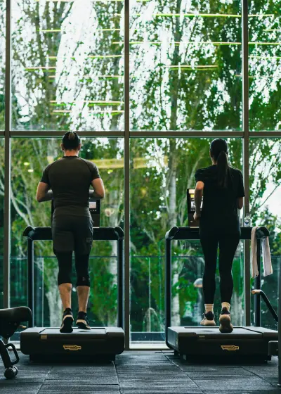 Man and woman running on treadmills facing large windows with a green forest outside.