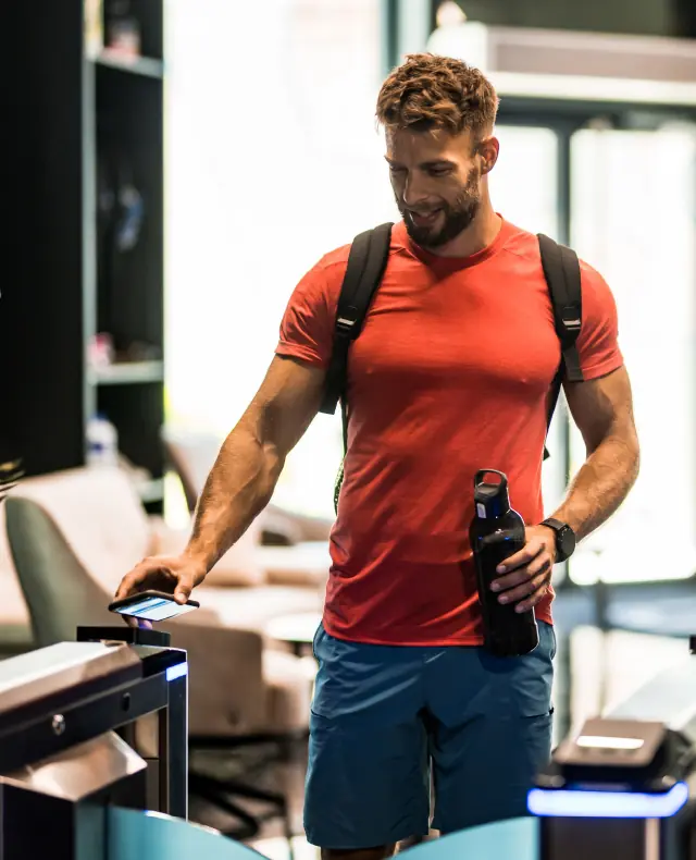 Man in red shirt and blue shorts using a smartphone to scan at a gym entrance while holding a water bottle.