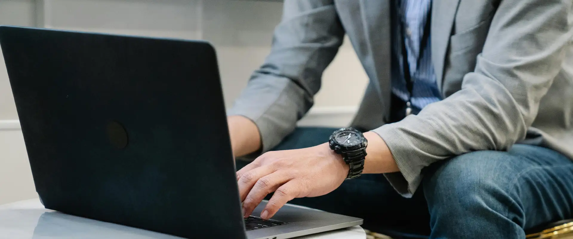 Person wearing a watch and blazer typing on a laptop placed on a round white table.