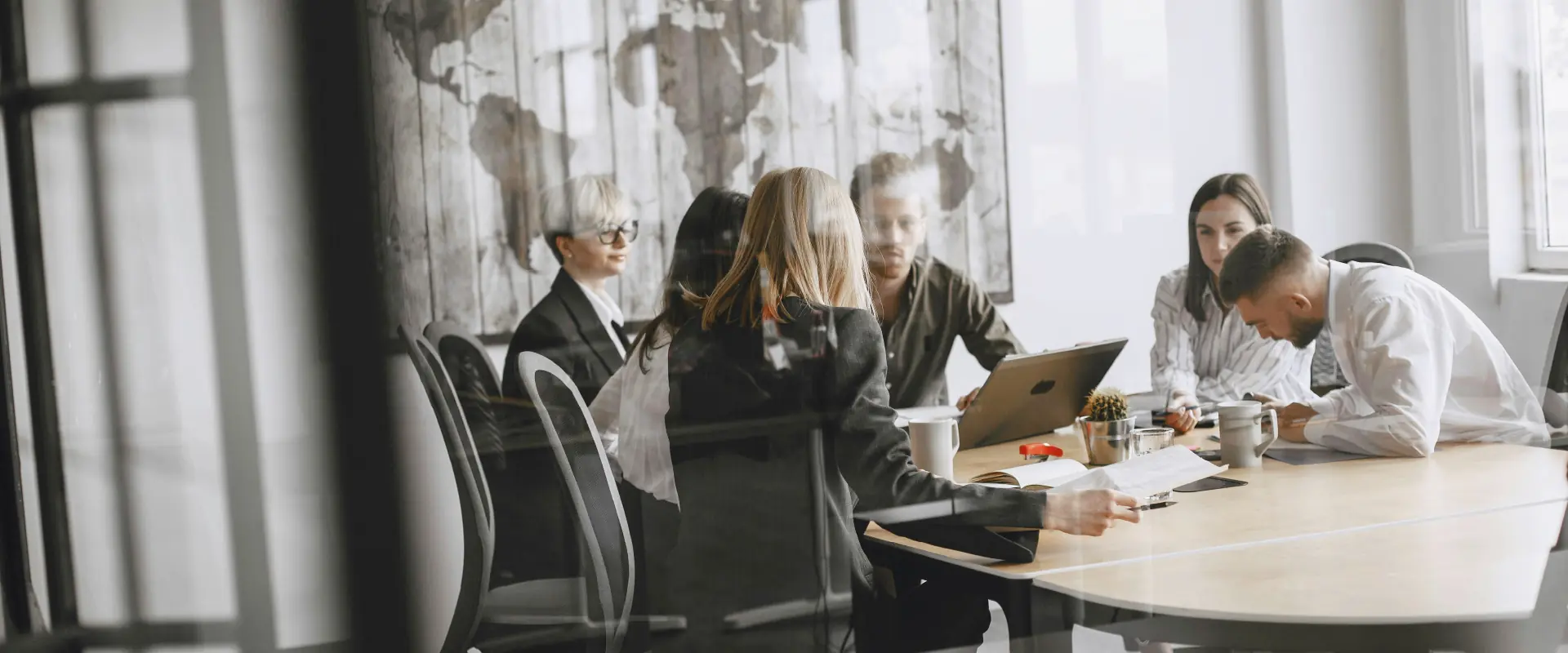 Five business people having a meeting around a table with laptops, documents, and coffee mugs in a bright office with a world map on the wall.
