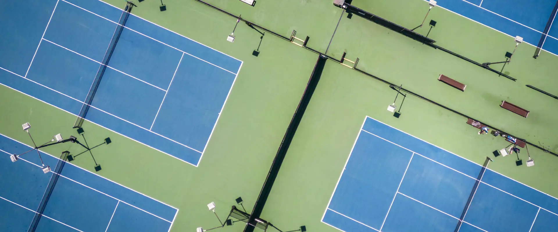 Aerial view of multiple blue and green outdoor tennis courts with benches and light poles.
