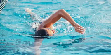 Person swimming freestyle in a clear blue pool wearing swim goggles and cap.