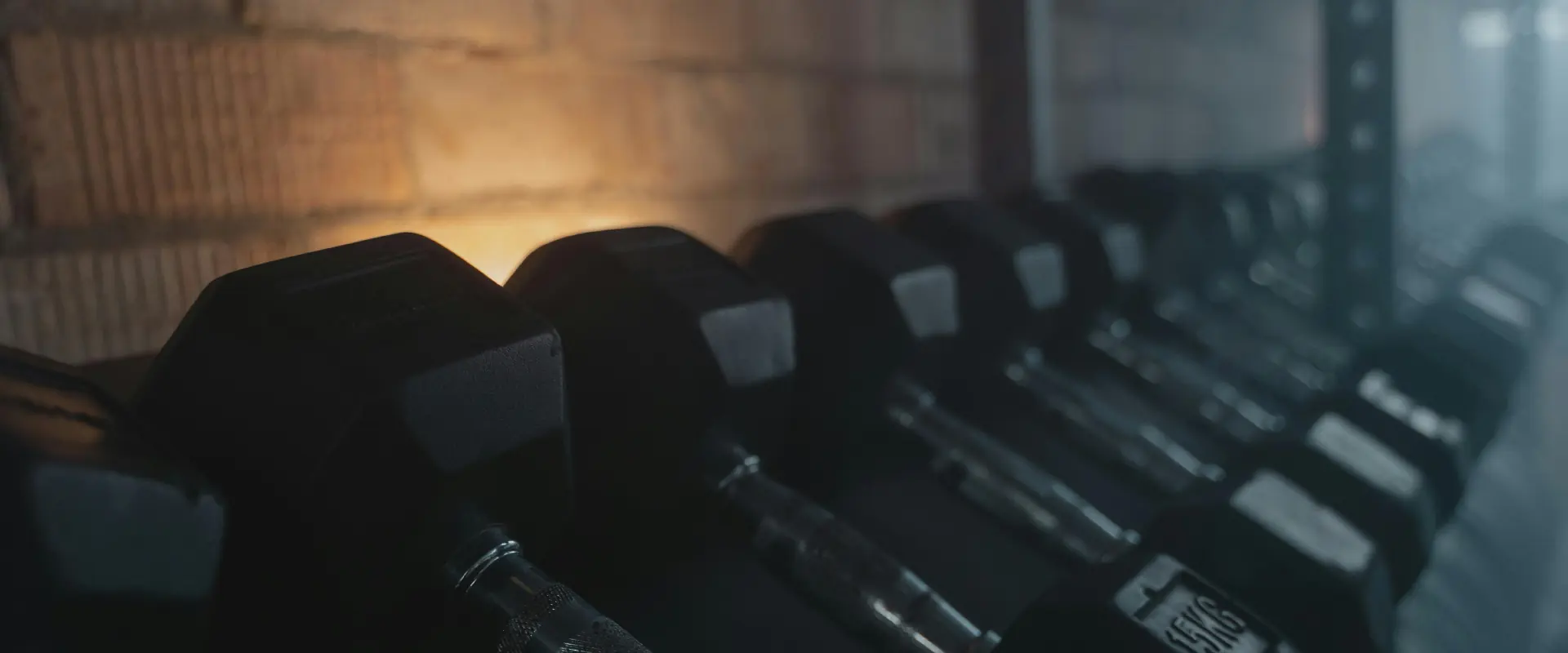 Row of black hexagonal dumbbells on a rack in a gym with a brick wall background.